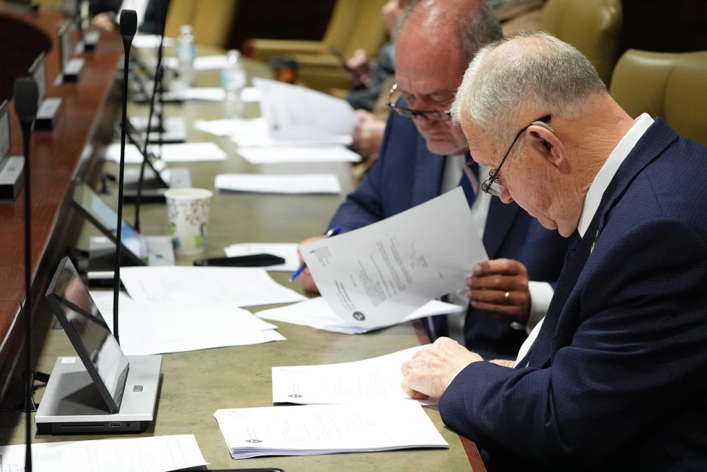 Senator Wallace studies a printed report in the foreground. Senator Hickey is turning pages in the report in the background.