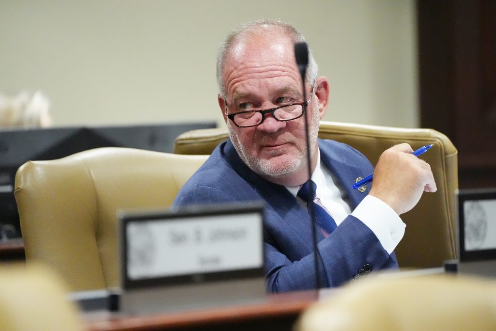 Senator Jimmy Hickey, Jr., looks over his glasses at a colleague during a committee meeting. He is seated and holding a blue pen in his hand.