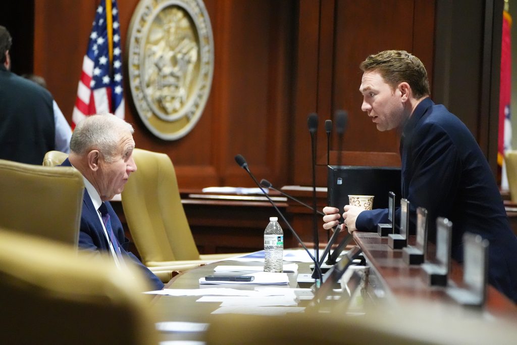 Senator Wallace is seated at one of the desks in a committee room. Senator Dees is leaning over the ledge talking with him. The American flag and Great Seal of the State of Arkansas are seen on the wall behind them.