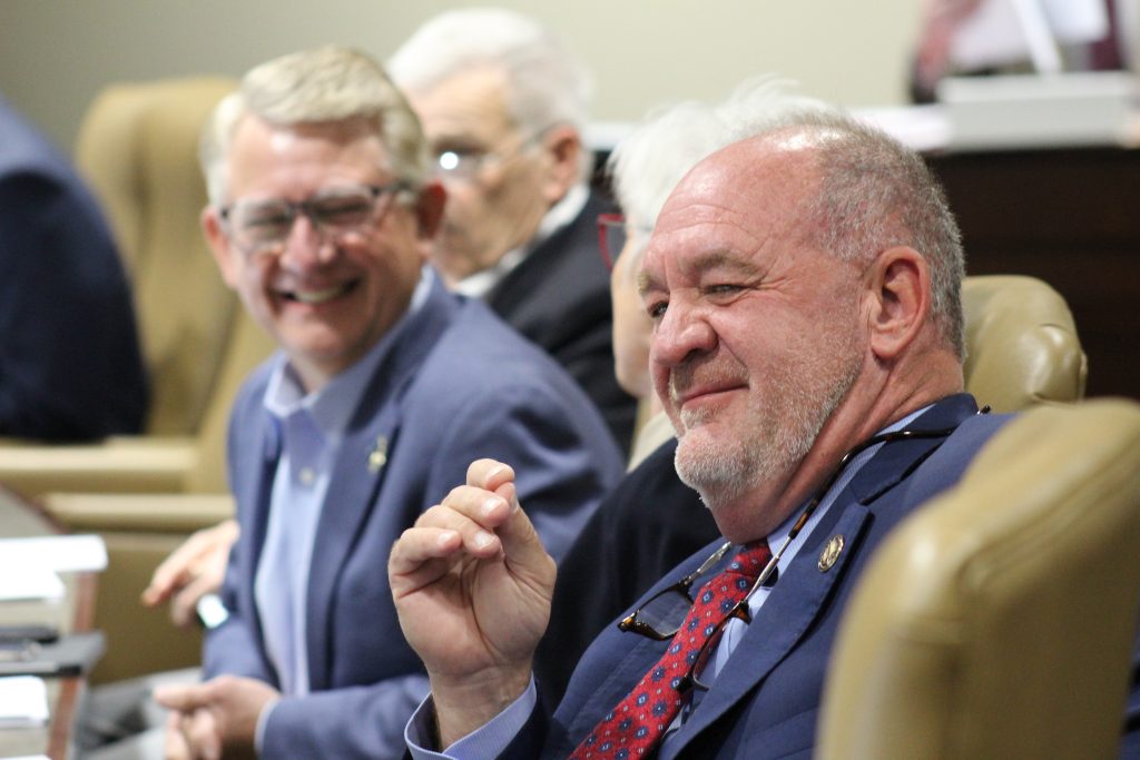 Senator Hickey smiles during a committee meeting. Senator Hill smiles in the background.
