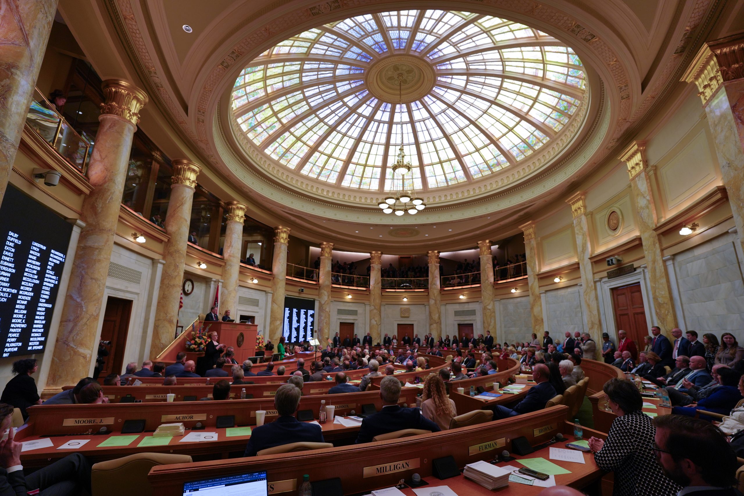 Wide angle photo of the Arkansas House of Representatives chamber. The photo shows from the stained glass dome down to the crowd below. Governor Sarah Sanders is speaking from the lectern.