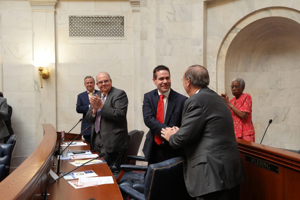 Senator Brad C. Simon shakes hands with Senator Mark Johnson as other colleagues applaud.