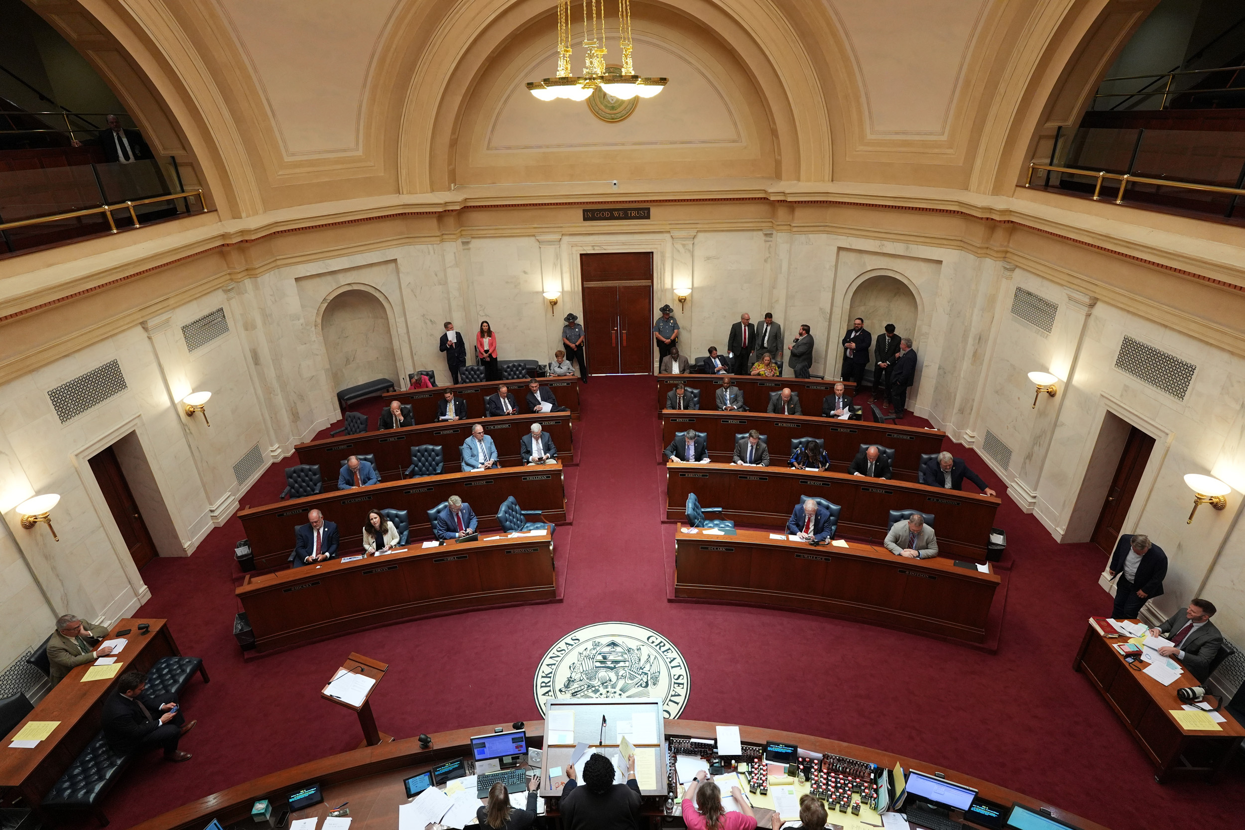 Shot of the full Senate chamber, taken from above. Senators are at their desks or in various places around the room.