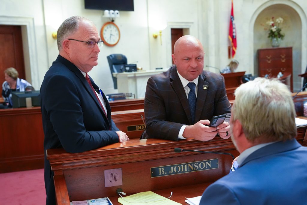 Senators Stone and McKee lean on Blake Johnson's desk as they talk to him. The front of the Senate chamber can be seen in the background.