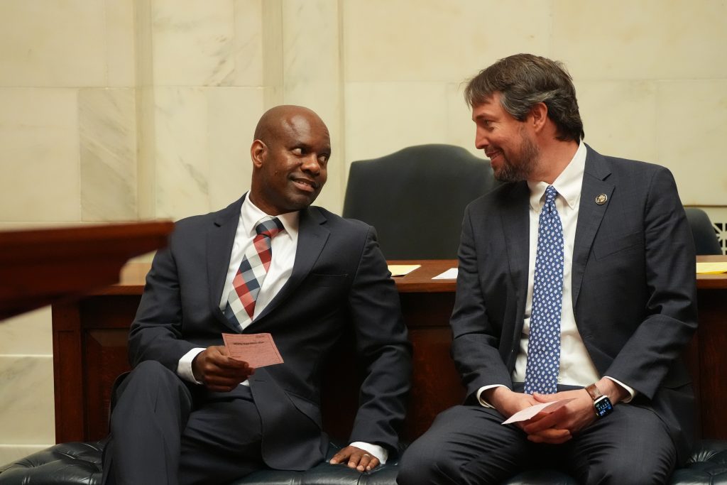 Senators Fred Love and Clarke Tucker talk on a bench in the Well in the Senate chamber.