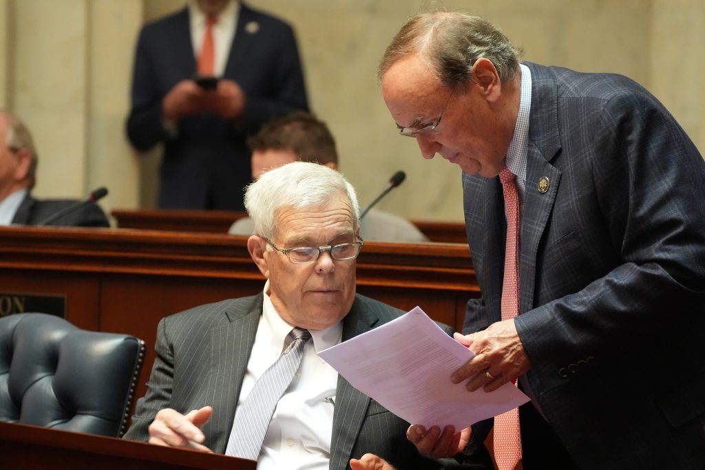 Senator Mark Johnson shows some papers to Senator Sullivan, who is seated at his desk in the Senate chamber.