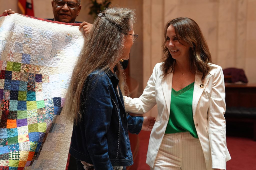 Senator Irvin congratulates a woman for her Arkansas quilt, seen partially in the background. They are standing in the Senate chamber.