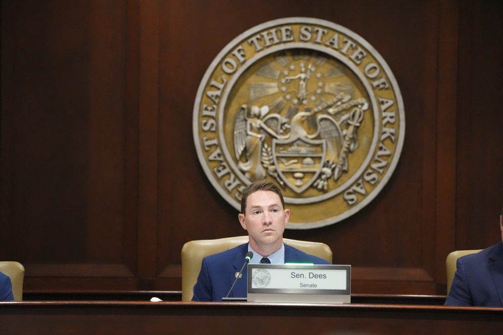 Senator Dees listens in a committee room. The Great Seal of Arkansas is on the wall behind him.