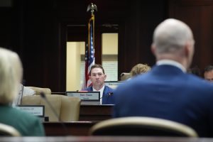 Senator Dees listens to testimony in a committee room. He is in focus in the background while the people testifying are in the foreground, out of focus.