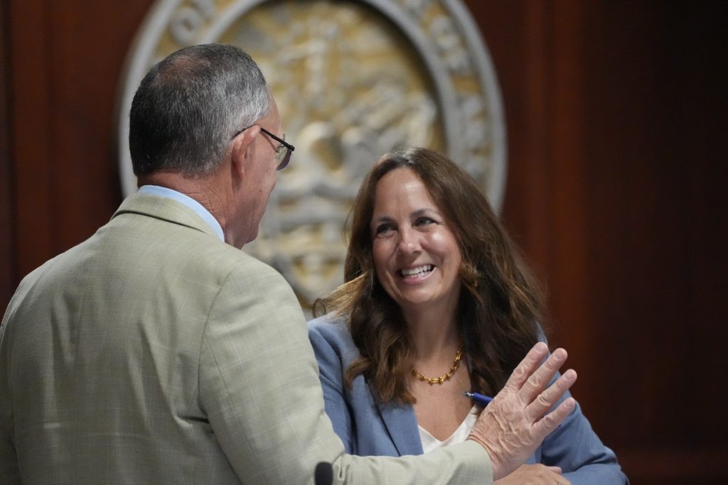 Senator Irvin smiles while talking with Senator Stone, who is seen from behind. They are talking in a committee room after a meeting.