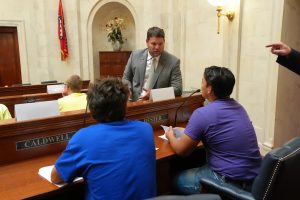 Senator Hester speaks with two Boys State delegates. The boys are seated at Senate desks and Senator Hester is standing in front of them gesturing as he talks.