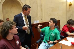 Senator Simon talks with a Boys State delegate in the Senate chamber. The boys are seated at the senate desks and Senator Simon is standing, holding a coffee cup.