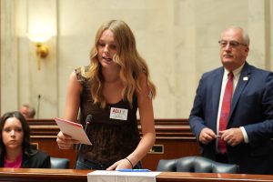 A young woman speaks in the Senate chamber. Senator Hammer is seen in the background.