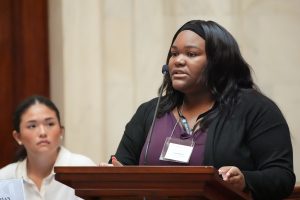A Girls State delegate speaks from the Well in the Senate chamber while another girl looks on in the background.